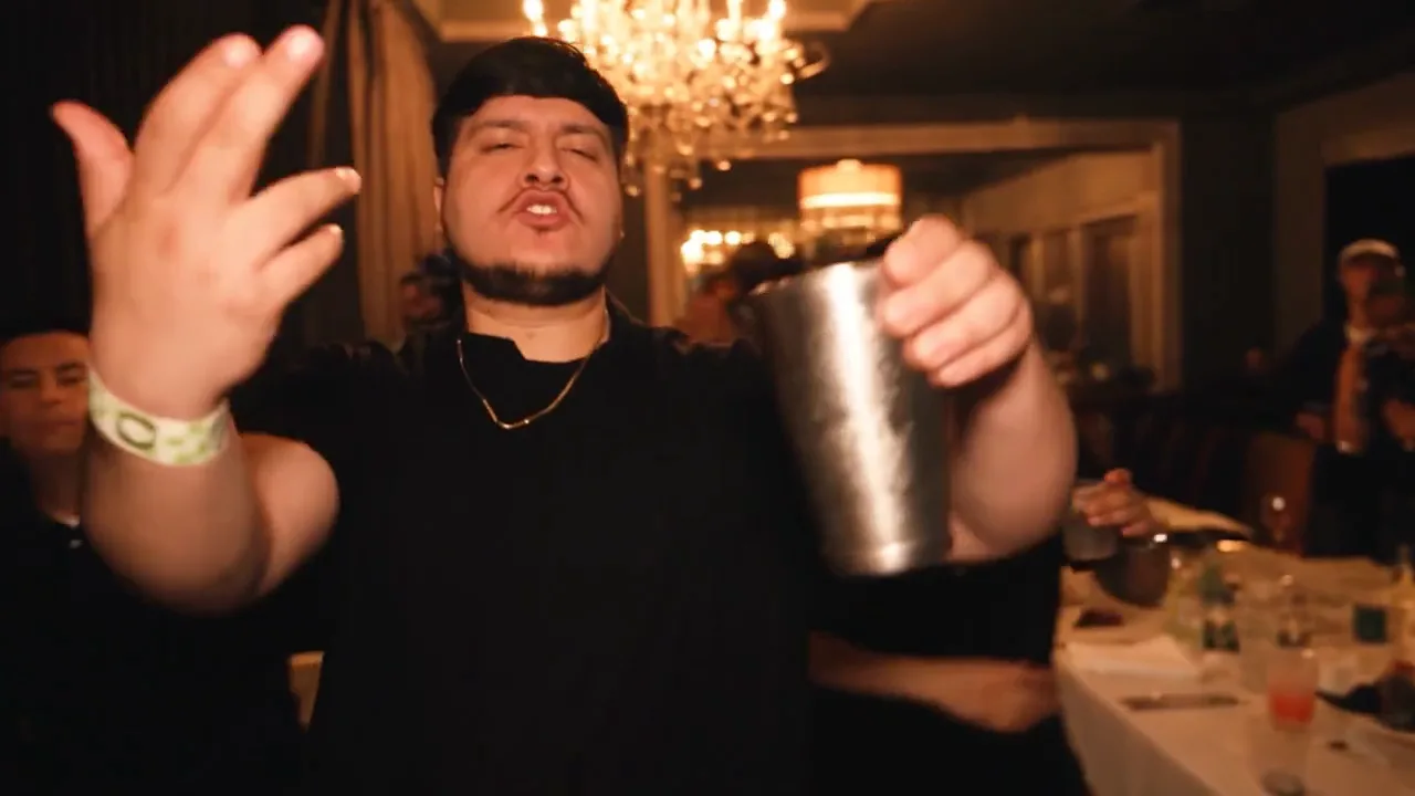 Original MV frame: performer close to the camera with an open hand and silver cup beneath a chandelier