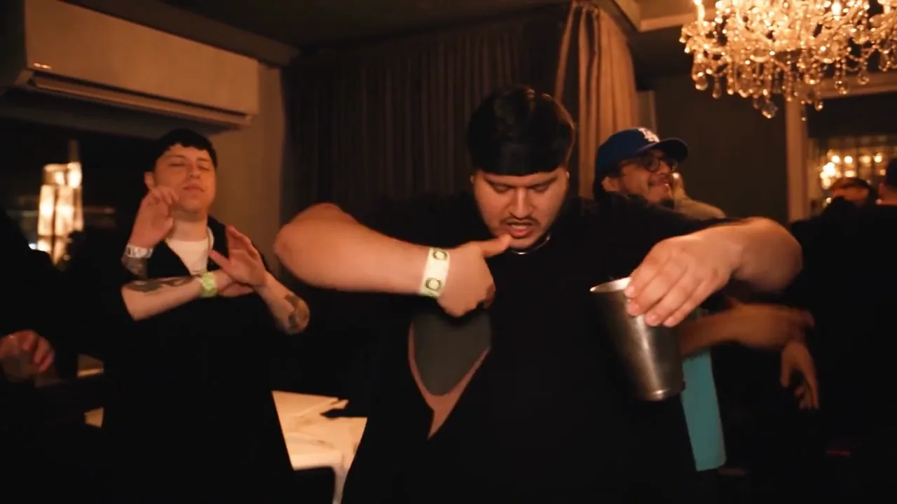 Original MV frame at nine seconds: performer leaning forward and pointing toward his chest while holding a cup